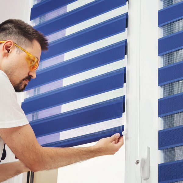 Male builder carpenter pulling down a blue jalousie on a window. Repairing and inspection fabric curtains. Duo system day and night blind. Interior decor element.
